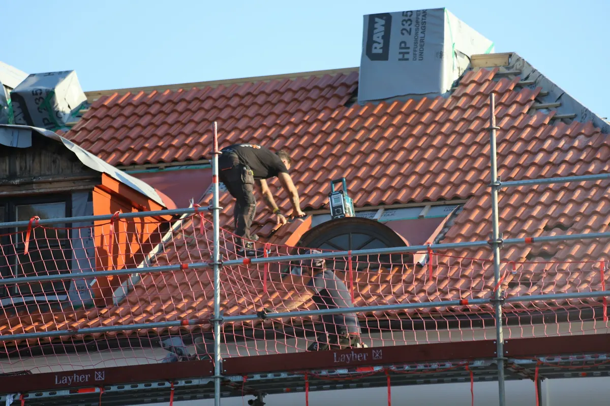 Dachdecker auf dem Gerüst montiert eine Dachziegelreihe an einer Gaube mit rundem Fenster