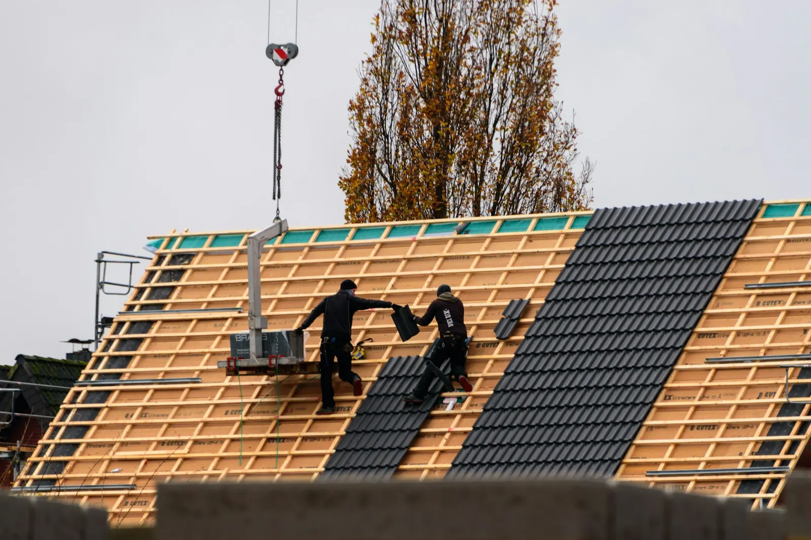 Zwei Dachdecker decken bei einem Neubau in Offenbach die Dachlatten mit Ziegeln ein, im Hintergrund ein Baukran